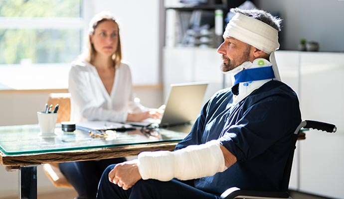 A man in a wheelchair wearing a head bandage, cervical neck brace, and arm cast, meeting with a professional woman