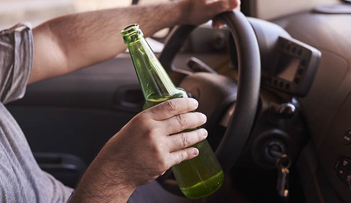 A driver's hands, one on the steering wheel and the other holding an open green glass bottle