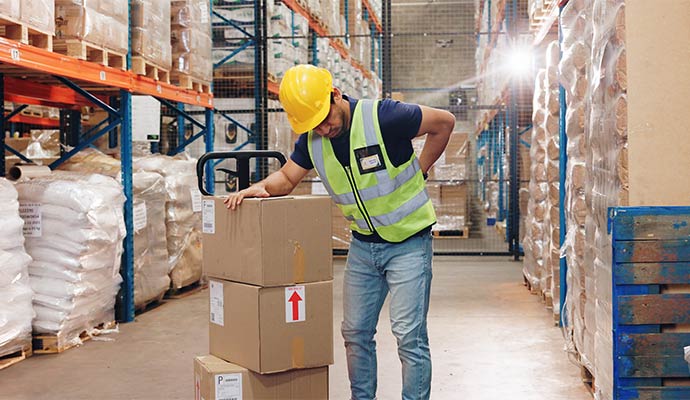 A warehouse worker is leaning against a stack of boxes while holding his lower back